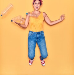 Young beautiful woman with pink short hair smiling happy. Jumping with smile on face around plastic bottles over isolated yellow background