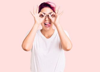 Young beautiful woman with pink hair wearing casual clothes and glasses doing ok gesture like binoculars sticking tongue out, eyes looking through fingers. crazy expression.