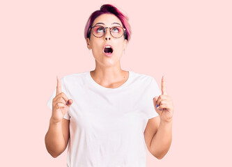 Young beautiful woman with pink hair wearing casual clothes and glasses amazed and surprised looking up and pointing with fingers and raised arms.