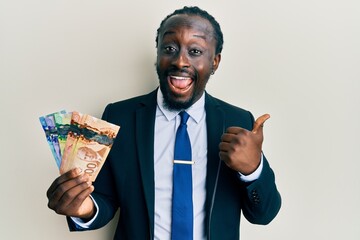 Handsome young black man wearing business suit and tie holding canadian dollars pointing thumb up...