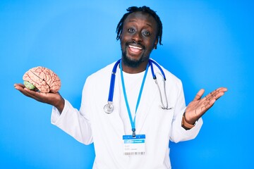 Young african american man with braids wearing doctor coat holding brain celebrating achievement...