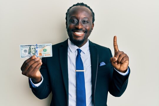 Handsome young black man wearing business suit and tie holding 100 dollars smiling with an idea or question pointing finger with happy face, number one