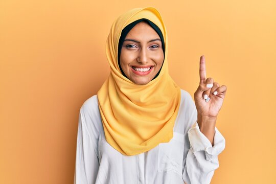 Young Brunette Arab Woman Wearing Traditional Islamic Hijab Scarf Showing And Pointing Up With Finger Number One While Smiling Confident And Happy.