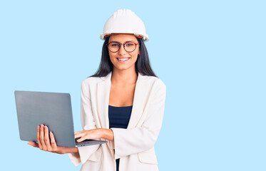 Young beautiful latin girl wearing architect hardhat holding laptop looking positive and happy standing and smiling with a confident smile showing teeth