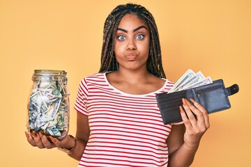 Young african american woman with braids with savings jar and wallet with dollars puffing cheeks with funny face. mouth inflated with air, catching air.