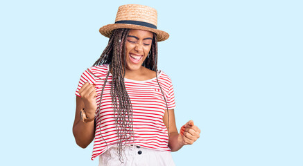 Obraz na płótnie Canvas Young african american woman with braids wearing summer hat celebrating surprised and amazed for success with arms raised and eyes closed. winner concept.