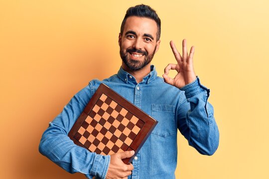 Young hispanic man holding chess doing ok sign with fingers, smiling friendly gesturing excellent symbol