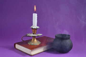 Candle in candlestick burning on an old book and witch smoky pot on the dark purple smoked background. Halloween concept