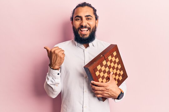 Young arab man holding chess pointing thumb up to the side smiling happy with open mouth