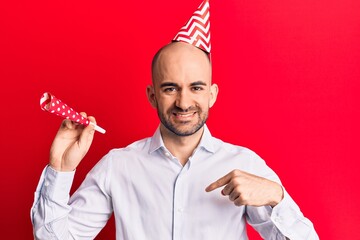 Young handsome bald man wearing party hat and trumpet pointing finger to one self smiling happy and...