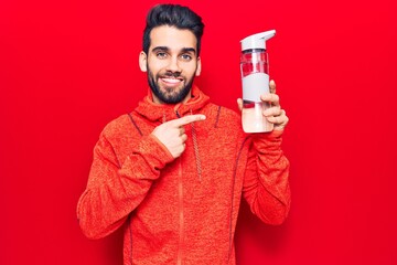 Young handsome man with beard wearing sportswear drinking bottle of water smiling happy pointing with hand and finger