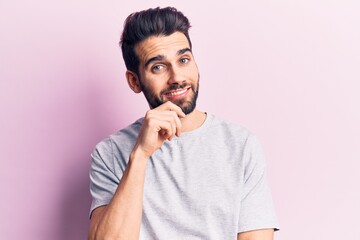 Young handsome man with beard wearing casual t-shirt smiling looking confident at the camera with crossed arms and hand on chin. thinking positive.