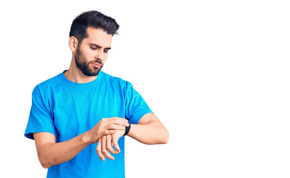 Young handsome man with beard wearing casual t-shirt checking the time on wrist watch, relaxed and confident