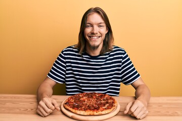 Handsome caucasian man with long hair eating tasty pepperoni pizza looking positive and happy standing and smiling with a confident smile showing teeth
