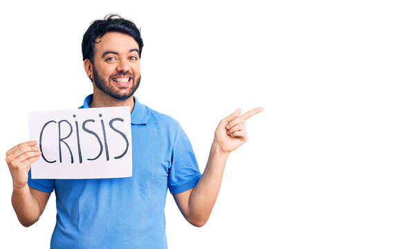 Young hispanic man holding crisis message paper smiling happy pointing with hand and finger to the side