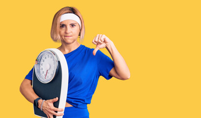 Young blonde woman wearing sportswear holding weighing machine with angry face, negative sign...
