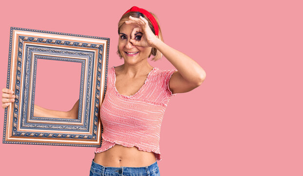 Young blonde woman holding empty frame smiling happy doing ok sign with hand on eye looking through fingers
