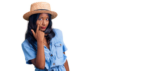 Young indian girl wearing summer hat pointing to the eye watching you gesture, suspicious expression