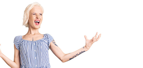 Fototapeta na wymiar Young blonde woman with tattoo wearing casual clothes crazy and mad shouting and yelling with aggressive expression and arms raised. frustration concept.