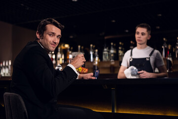 a handsome man poses at the bar with a cocktail in his hands against the background of the bartender and bottles.