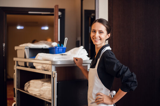 A smiling hotel maid with cleaning cart and cleaning supplies, looking at the camera and posing.