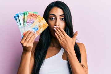 Beautiful hispanic woman holding swiss franc banknotes covering mouth with hand, shocked and afraid for mistake. surprised expression