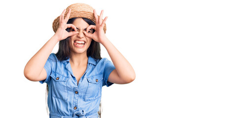 Brunette teenager girl wearing summer hat doing ok gesture like binoculars sticking tongue out, eyes looking through fingers. crazy expression.