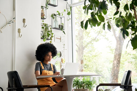 Happy stylish hipster African American female student with Afro hair using laptop computer sitting at table learning at home, in cozy modern cafe, remote studying, watching video, working in internet.