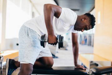 Side view of African American man in sportswear leaning on bench and doing row exercise with dumbbell during training in gym.