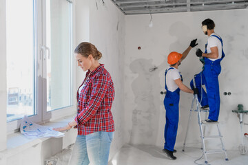 A customer stands by the window and examines the diagram of the house. Two builders drill a wall while standing on a stepladder