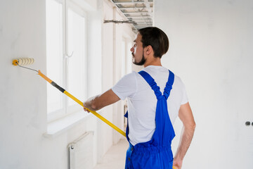 A bearded construction worker in a blue overalls is painting a wall in the hallway. He uses a roller