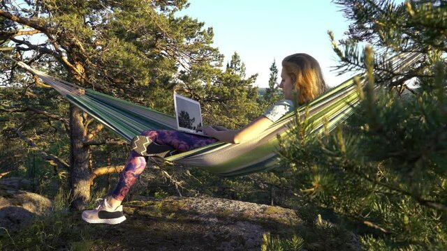 A young woman working on a laptop while lying in a hammock in the woods. Self-isolation, freelancing, remote work