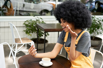 Fototapeta na wymiar Happy African American young hipster woman holding smart phone making video call, shooting social media blog talking, virtual meeting friend sitting at outdoor cafe table. Social distance concept.
