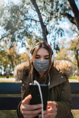 A young woman wearing face mask using her smartphone and listening music at park