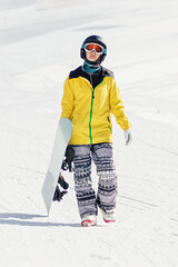 Fototapeta na wymiar Young woman with snowboard in hand walking on ski slope, she's looking at camera, copy space