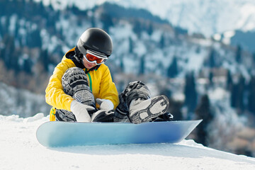 Fototapeta na wymiar Young woman snowboarder sits in the snow and fastens fastenings on a snowboard before the descent