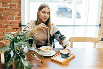 Young woman in casual outfit takes photos of food on smartphone in the cafe