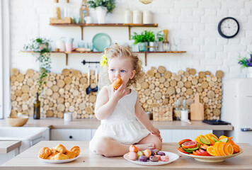 A little girl poses in the kitchen during Breakfast, eating delicious macaroons, cakes, cookies.