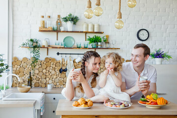 Family portrait of a happy mother, daughter and father posing in the kitchen during Breakfast, eating delicious macaroons, cakes, cookies. Married good relations, love each other