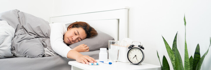 Obraz na płótnie Canvas sick Asian woman lying in bed take medicines pills and glass of water from bedside table to relieve symptoms.
