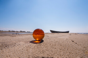 Fototapeta na wymiar Orange glass ball on sand near the sea. Abstract conceptual photo.