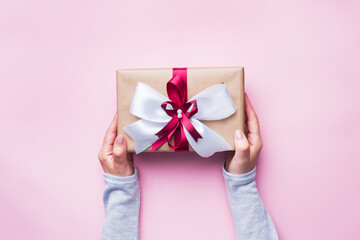 Gift or present box with a big bow in the hands of a woman on a pink table. Flatlay composition for Christmas birthday, mother day or wedding.
