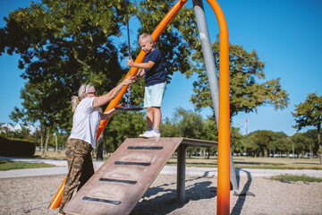 Young boy playing on a zip line in public park during summer day. He is with grandmother.