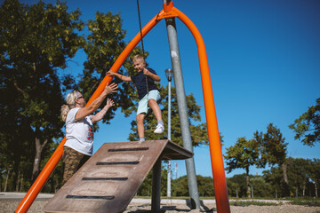 Young boy playing on a zip line in public park during summer day. He is with grandmother.