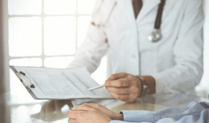 Unknown male doctor and woman-patient discussing current health examination while sitting in sunny clinic and using clipboard. Good medical service in hospital