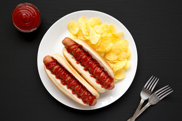 Tasty American Hot Dog with Potato Chips on a white plate on a black surface, top view. Flat lay, overhead, from above.