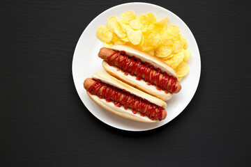 Tasty American Hot Dog with Potato Chips on a white plate on a black background, top view. Flat lay, overhead, from above. Copy space.