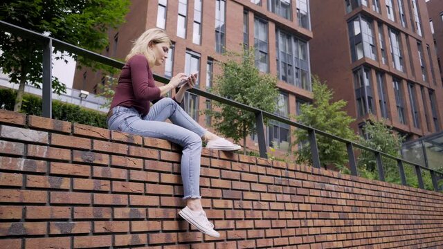 pretty blonde woman in stylish casual clothes surfs internet with modern smartphone sitting on red brick fence in city street on sunny summer day