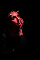 Halloween concept. Dreaming Demon woman with red face and small horns, wears black clothes poses against black background with copy space. Ideas for halloween body art