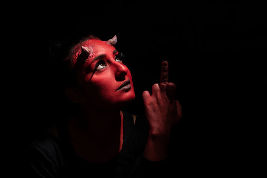 Halloween concept. Headshot of demon woman with red face makeup and small horns, wears black clothes, showing middle finger doing bad expression, poses against black background with copy space.